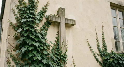 A plain stone cross mounted on an outside wall partly covered in ivy, photographed in soft overcast light.
