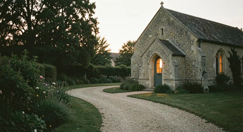 A gravel path curving through a garden toward a small stone chapel at dusk, warm window light, trees in silhouette.
