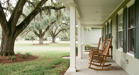 A covered porch with wooden rocking chairs looking over a lawn shaded by live oaks.